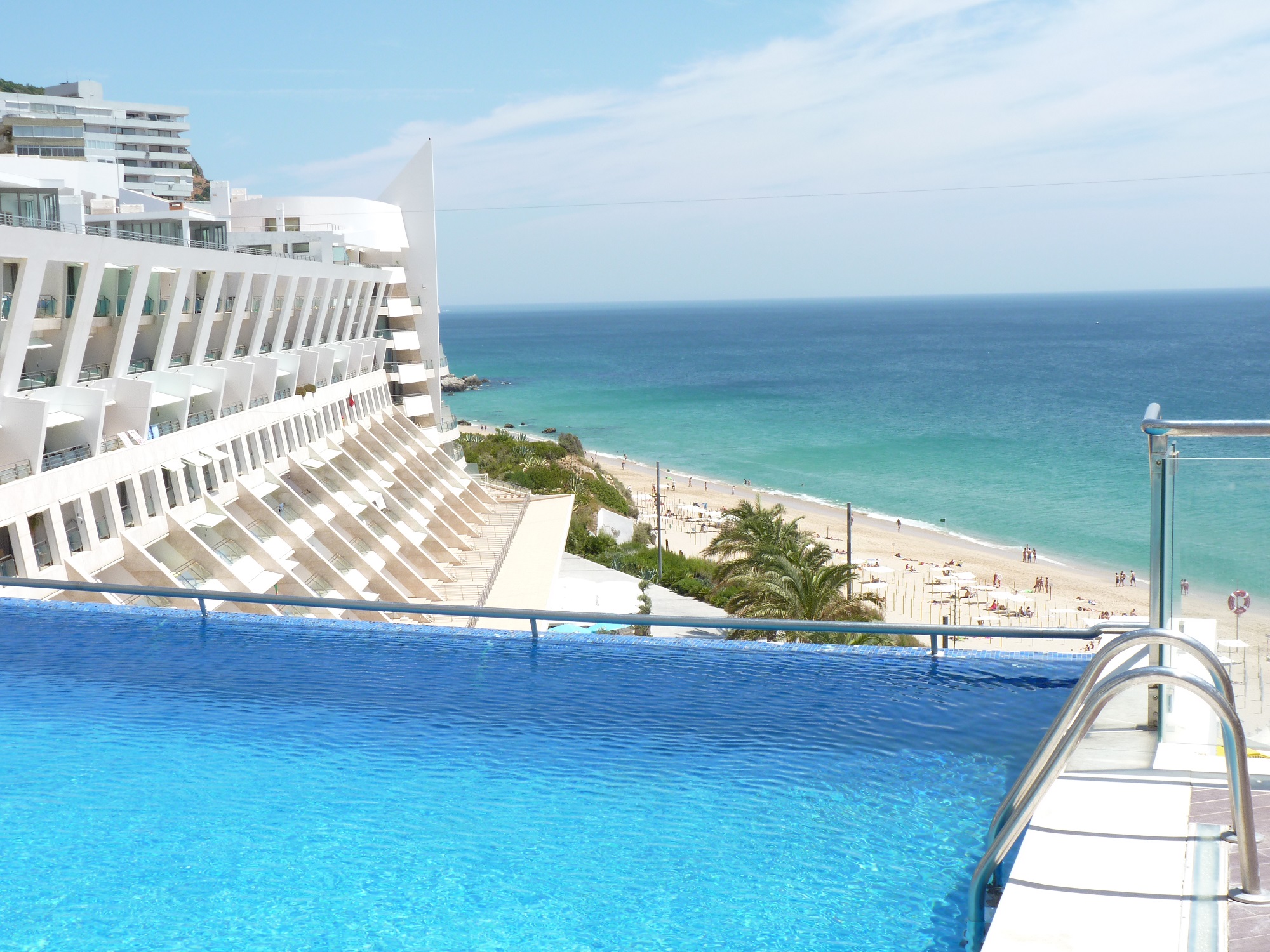 Sesimbra Hotel & SPA med utsikt över en pool, stranden och havet en solig dag.